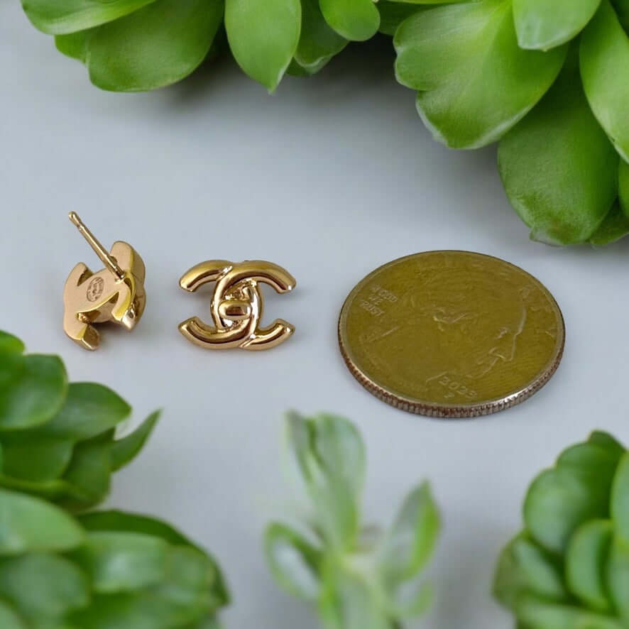 Chanel Gold CC Smaller Post Earrings next to a coin, showcased among greenery. Iconic Chanel Lock Style CC Earrings.