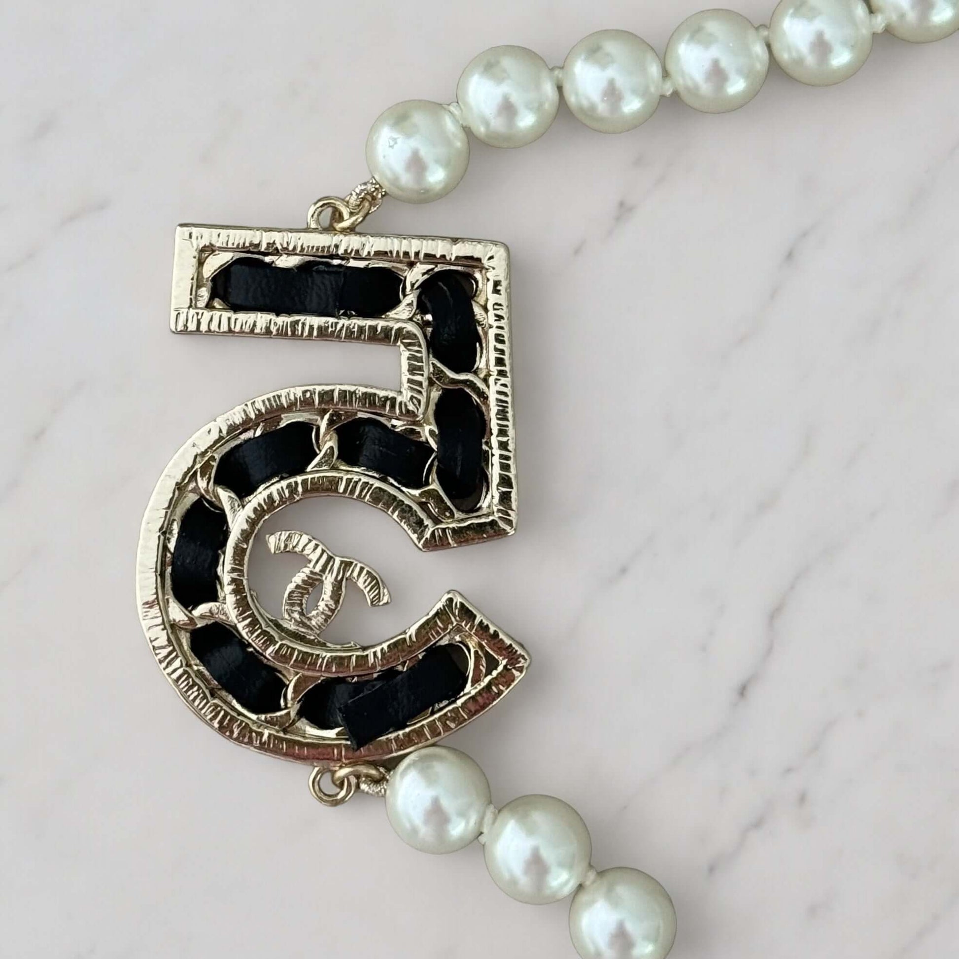 Chanel Knotted Pearl No.5 CC Sweater NecklaceIndulge in this gorgeous necklace adorned with a hand knotted pearl, Number 5 Chanel perfume charm, and sparkling crystals. The playful CC logo adds a touch of gold to the elegant black leather. Perfect for add