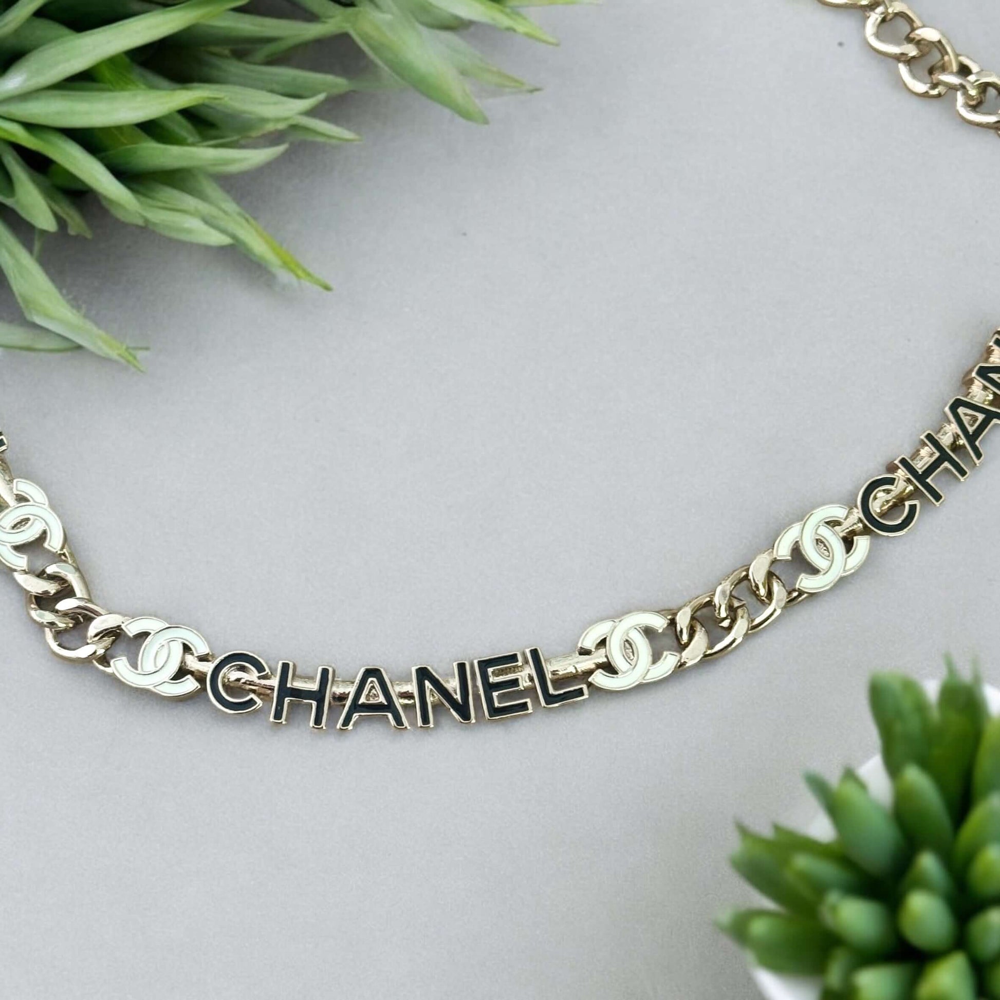 Chanel Strass Chain Adjustable Choker with black enamel CC logos on gold chain amidst green plants.