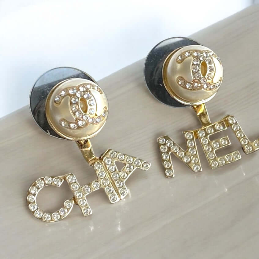 Chanel Gold CC Crystals Enamel Earrings featuring pearlish off-white enamel center and crystal-encrusted "CHA" and "NEL" wording in excellent pre-owned condition.