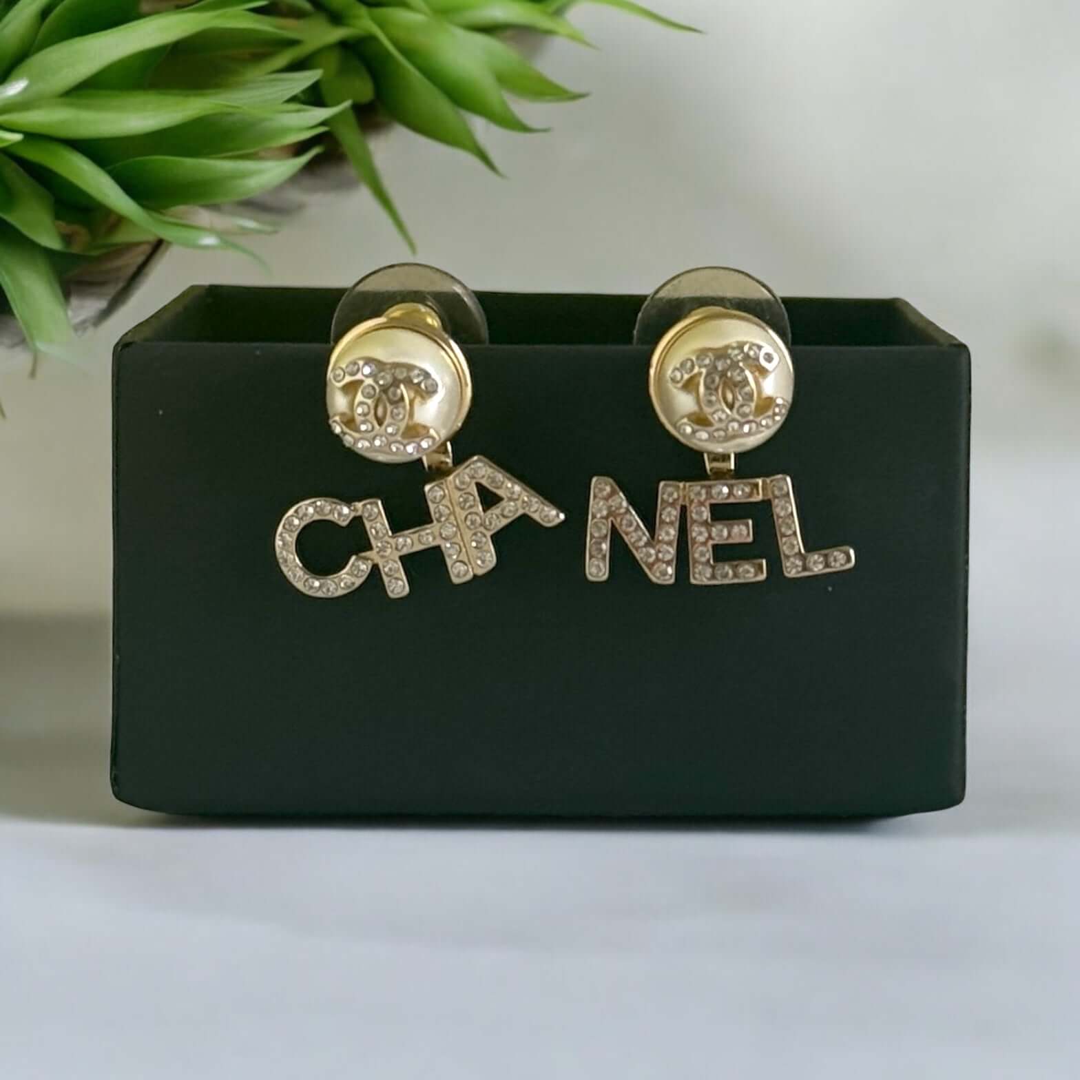 Chanel Gold CC Crystals Enamel Drop Earrings with "CHA" and "NEL" design, off-white enamel, clear crystals, on black display.