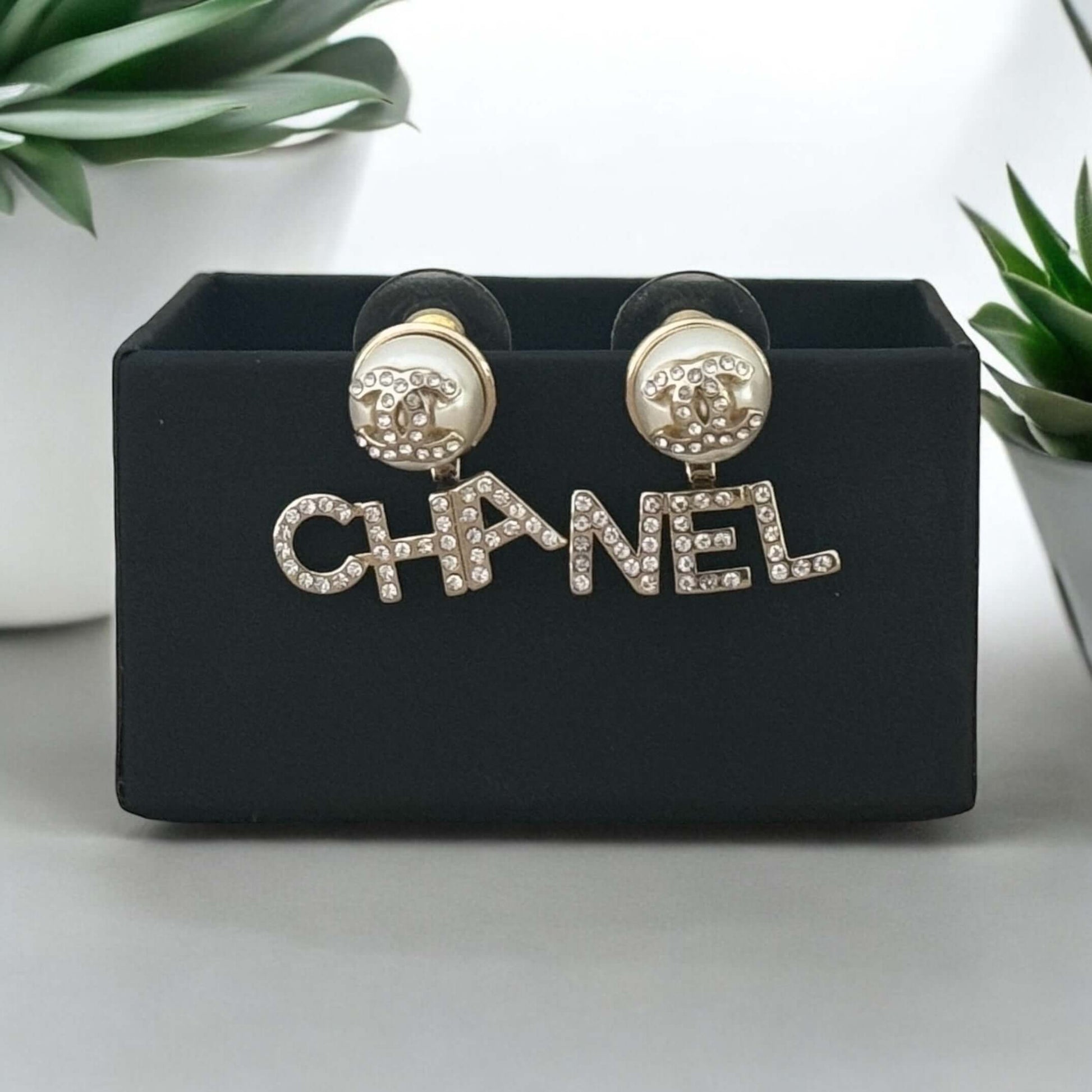 Chanel iconic gold earrings with clear crystals, off-white enamel CC center, "CHA" and "NEL" on each side, on display with gift box.