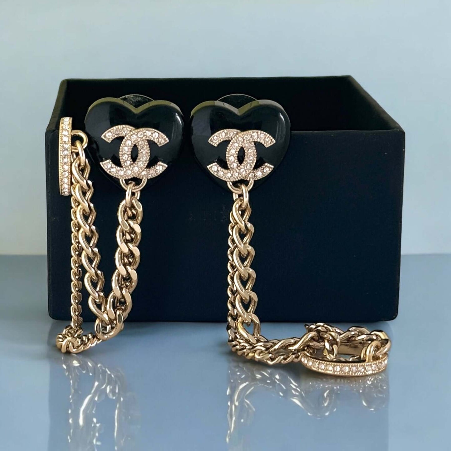 Chanel CC gold and black chain heart earrings with ear cuff, featuring crystals and enamel details, in front of Chanel box.